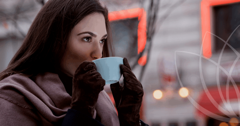 Naine joob sooja jooki helesinisest tassist, kandes pruune kindaid ja lillat salli, taustal hägused linna tuled