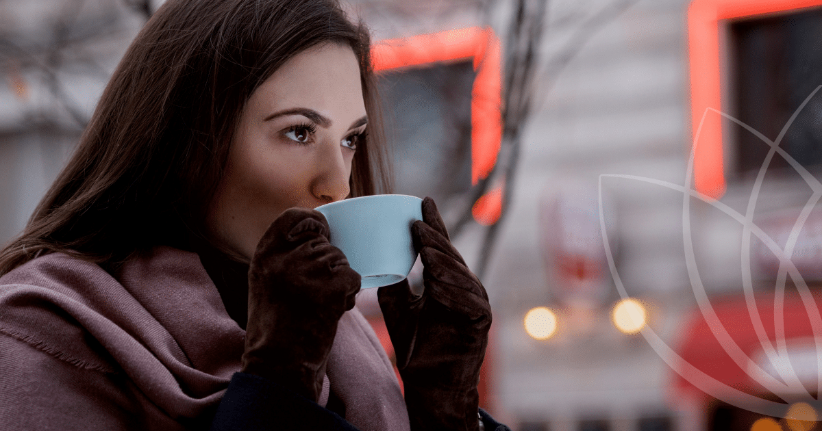Naine joob sooja jooki helesinisest tassist, kandes pruune kindaid ja lillat salli, taustal hägused linna tuled