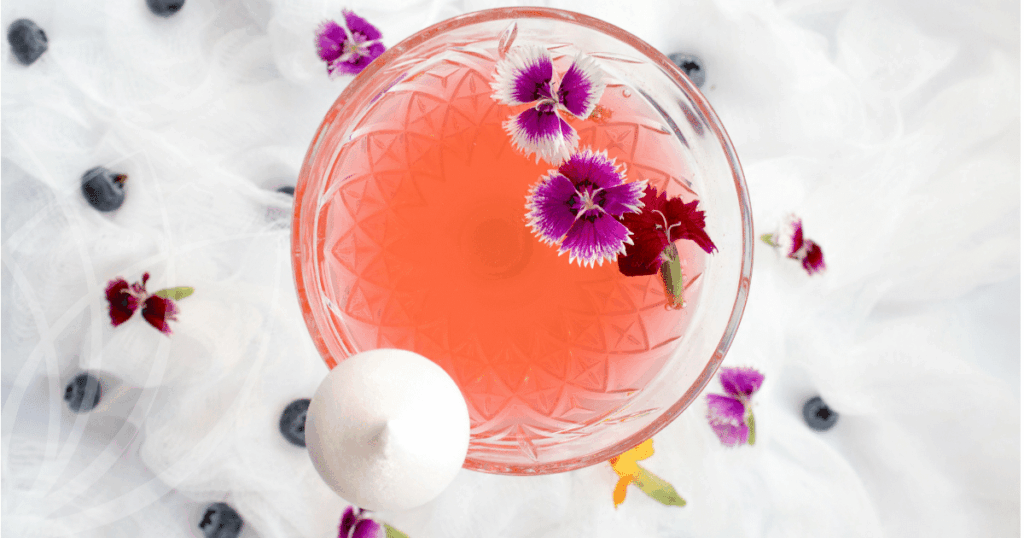 Klaasist kokteiliklaasis roosa joogi, valgete ja lillade õite ning mustikatega, taustaks valge pitsiline kangas.