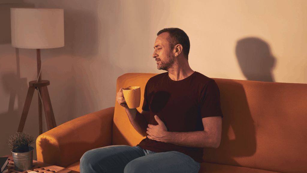 Mees istub diivanil, käes teetass, hoiab kõhust kinni, näol grimmass