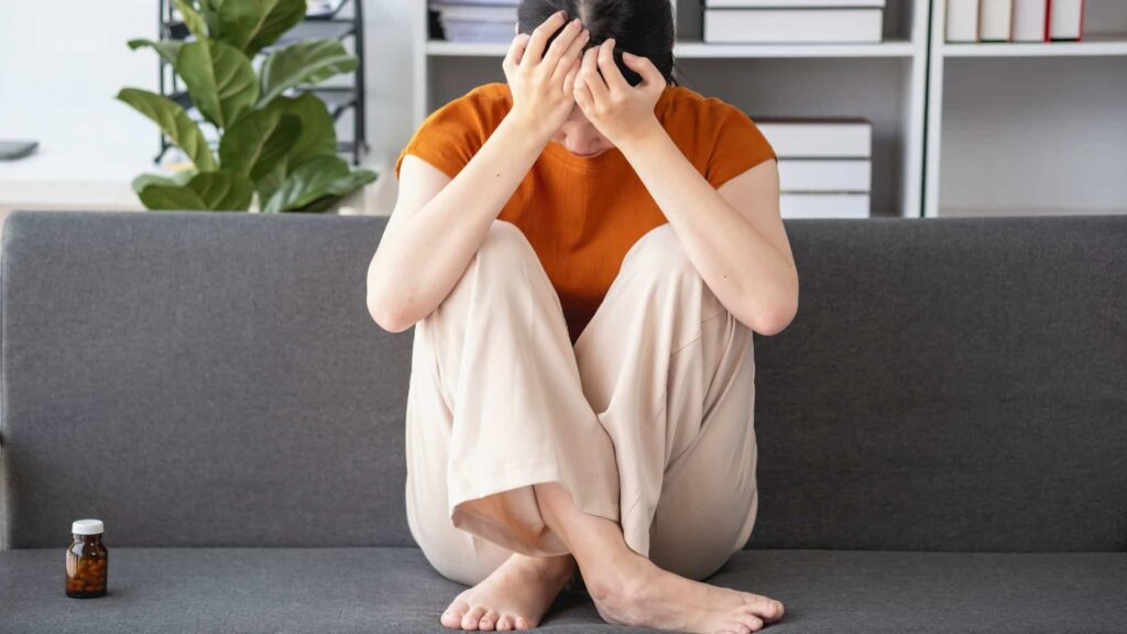 Naine istub diivanil ja hoiab kätega peast kinni, kõrval ravimipurk